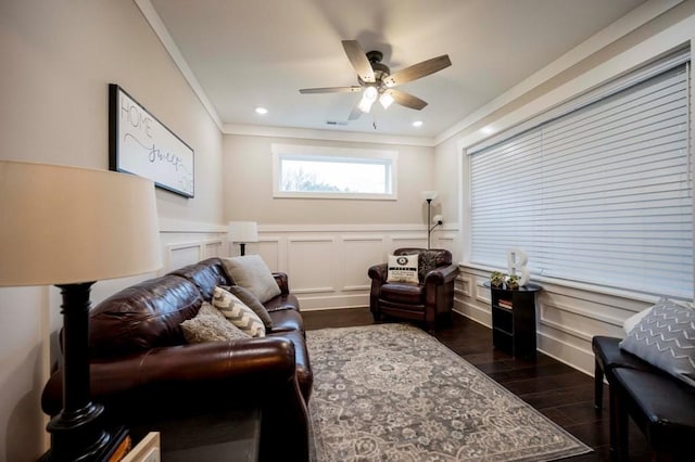 living area featuring a ceiling fan, ornamental molding, dark wood-type flooring, wainscoting, and a decorative wall