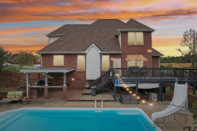 pool at dusk with a patio, a wooden deck, exterior kitchen, and a water slide