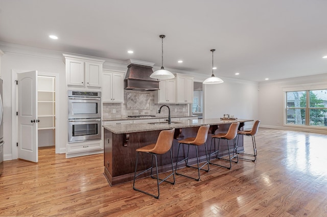 kitchen featuring stainless steel appliances, light stone counters, a kitchen island with sink, white cabinets, and custom range hood