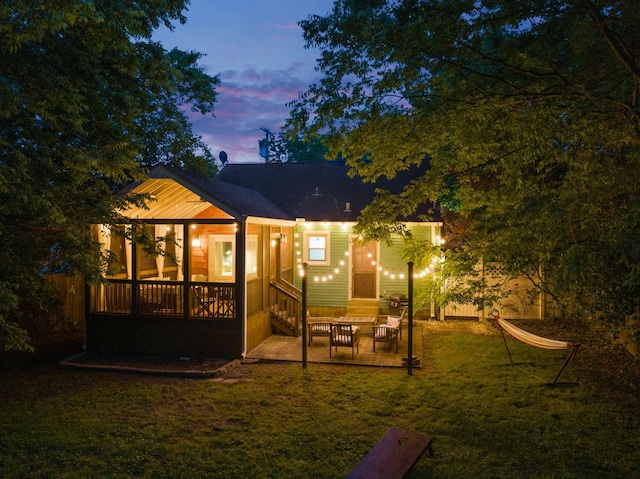 back house at dusk featuring a patio, a sunroom, and a lawn