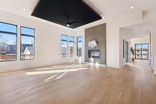 unfurnished living room featuring ceiling fan, a large fireplace, light wood-type flooring, and a tray ceiling