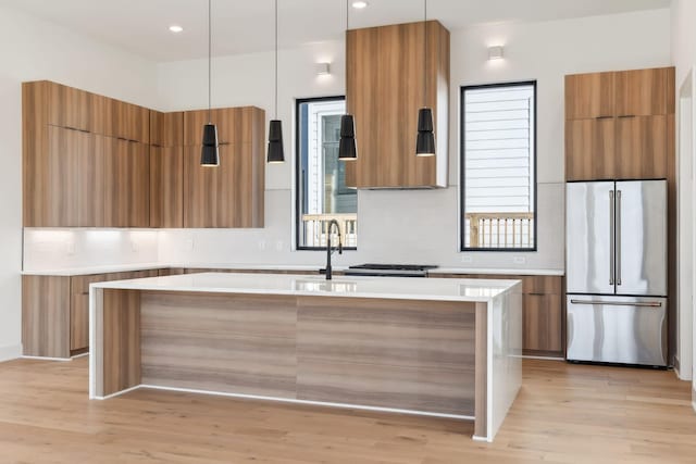 kitchen featuring hanging light fixtures, an island with sink, light hardwood / wood-style floors, decorative backsplash, and stainless steel built in fridge