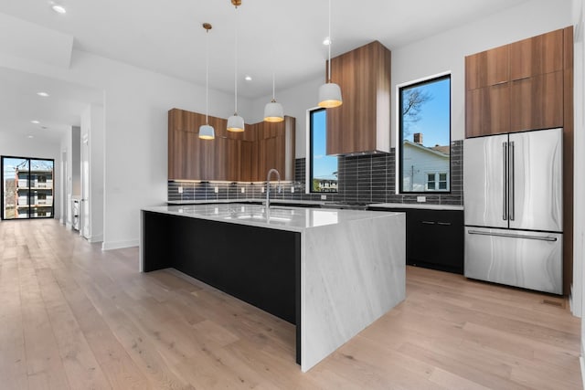 kitchen featuring decorative backsplash, light wood-type flooring, decorative light fixtures, a center island with sink, and stainless steel refrigerator