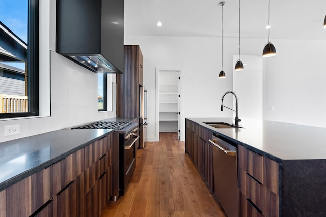 kitchen with wall chimney range hood, sink, appliances with stainless steel finishes, decorative light fixtures, and wood-type flooring