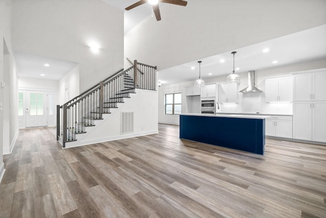 kitchen featuring light wood-type flooring, wall chimney exhaust hood, a kitchen island with sink, decorative light fixtures, and white cabinetry