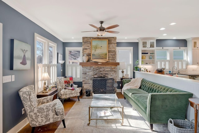 living room with a stone fireplace, sink, light wood-type flooring, ceiling fan, and crown molding