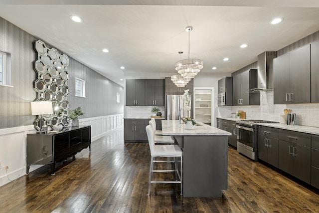 kitchen with stainless steel appliances, a kitchen island with sink, wall chimney range hood, dark hardwood / wood-style floors, and hanging light fixtures