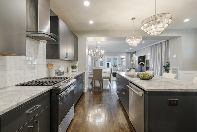 kitchen with hanging light fixtures, stainless steel appliances, wall chimney range hood, an inviting chandelier, and an island with sink