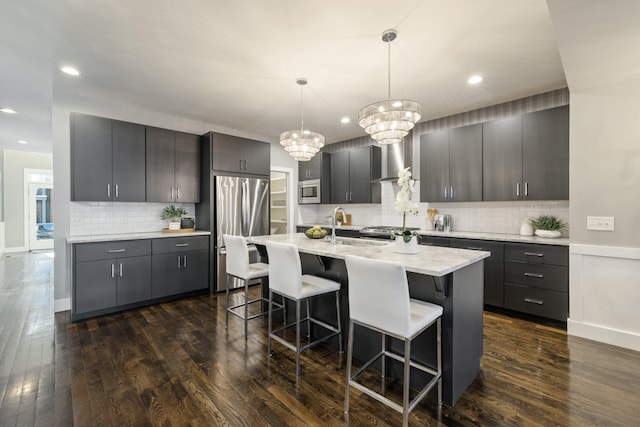 kitchen with dark hardwood / wood-style flooring, stainless steel appliances, wall chimney range hood, decorative light fixtures, and a center island with sink