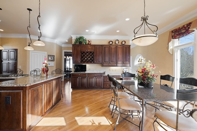 kitchen featuring light hardwood / wood-style flooring, hanging light fixtures, light stone counters, ornamental molding, and an island with sink