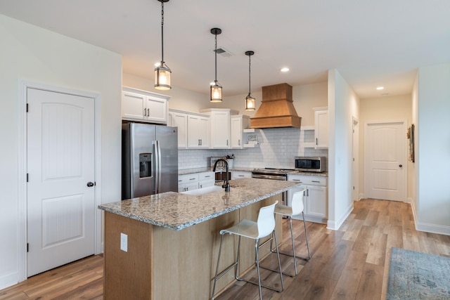 kitchen with white cabinets, a center island with sink, decorative light fixtures, custom range hood, and stainless steel appliances