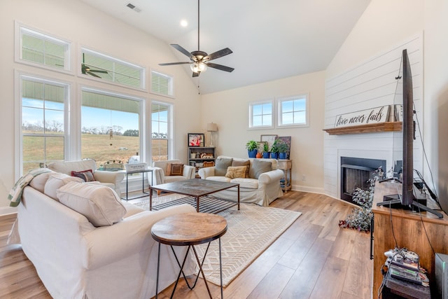 living room with a large fireplace, ceiling fan, light hardwood / wood-style flooring, and a towering ceiling