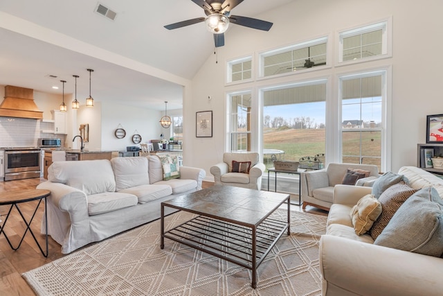 living room with ceiling fan with notable chandelier, light wood-type flooring, sink, and high vaulted ceiling