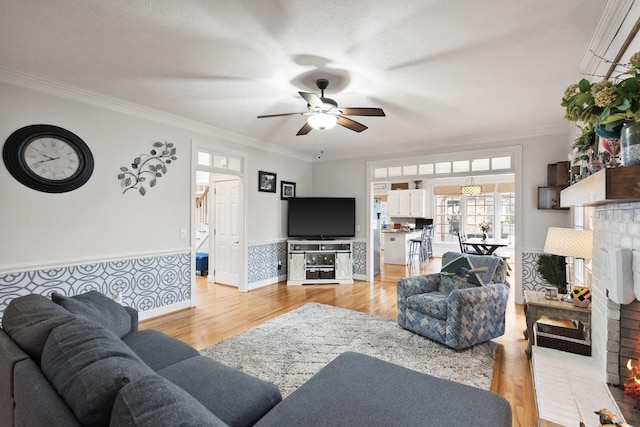 living room featuring light wood-type flooring, crown molding, ceiling fan, a brick fireplace, and a textured ceiling