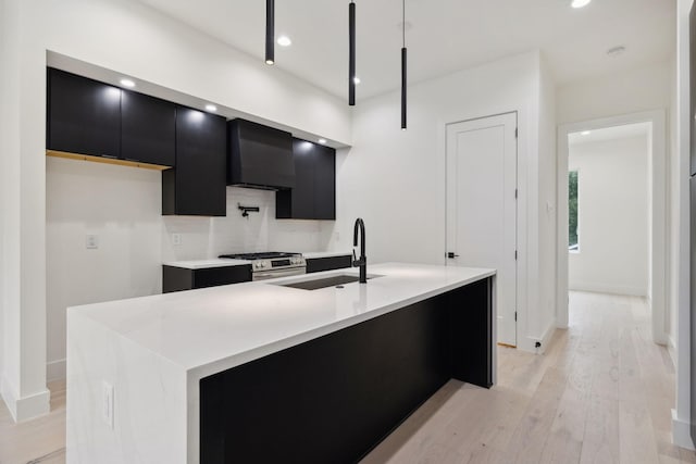 kitchen featuring light wood-type flooring, wall chimney exhaust hood, stainless steel range, sink, and an island with sink