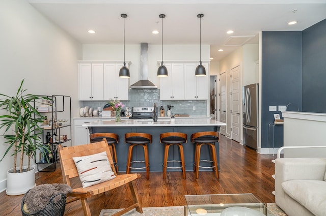 kitchen with stainless steel appliances, a kitchen island with sink, hanging light fixtures, and wall chimney range hood