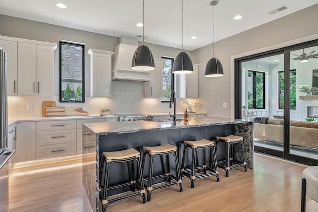 kitchen with white cabinets, custom range hood, an island with sink, and light hardwood / wood-style flooring