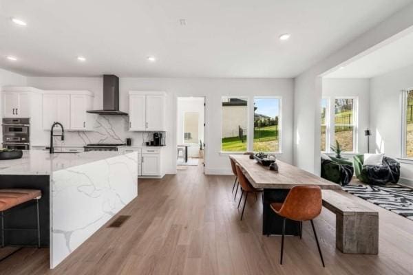 kitchen featuring light stone counters, double oven, sink, wall chimney range hood, and white cabinetry