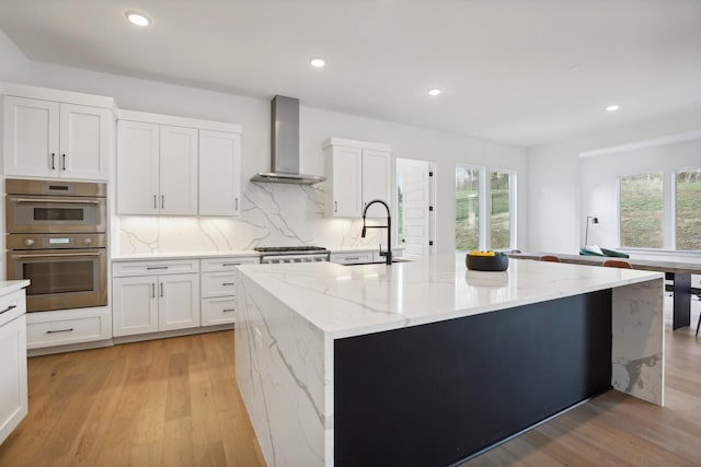 kitchen with sink, wall chimney range hood, light stone counters, a kitchen island with sink, and white cabinets