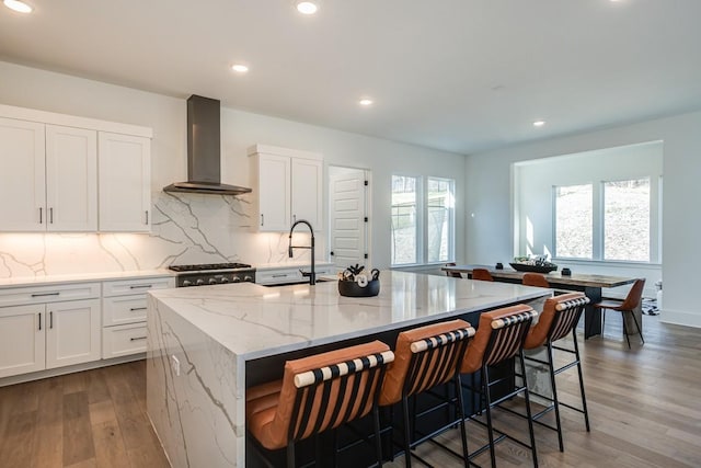 kitchen featuring white cabinets, wall chimney exhaust hood, light stone counters, and a kitchen island with sink