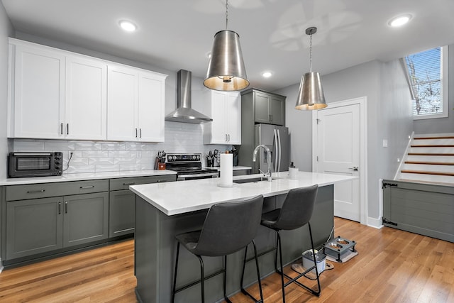 kitchen featuring wall chimney exhaust hood, stainless steel appliances, an island with sink, decorative light fixtures, and gray cabinets