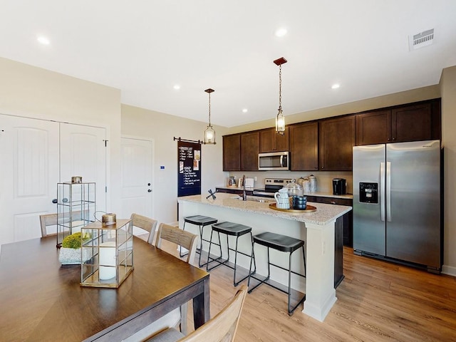 kitchen featuring dark brown cabinetry, light stone countertops, stainless steel appliances, light hardwood / wood-style floors, and a center island with sink