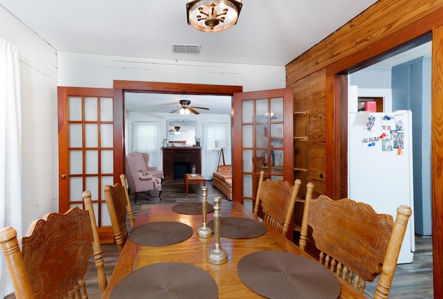 dining room featuring wooden walls, french doors, ceiling fan, and a brick fireplace