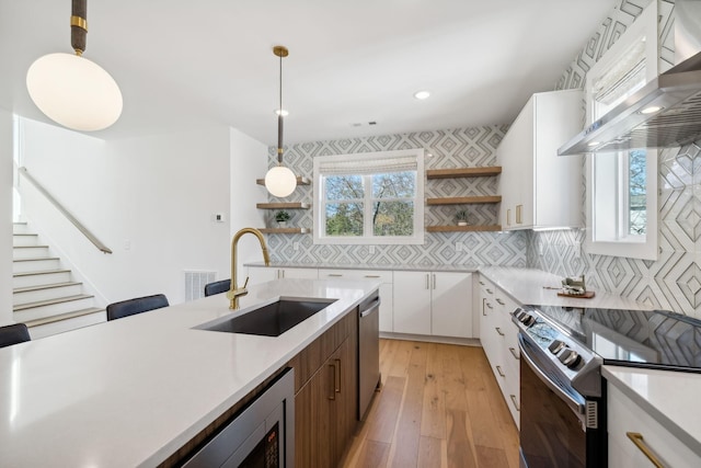 kitchen with white cabinets, wall chimney range hood, sink, decorative light fixtures, and stainless steel appliances