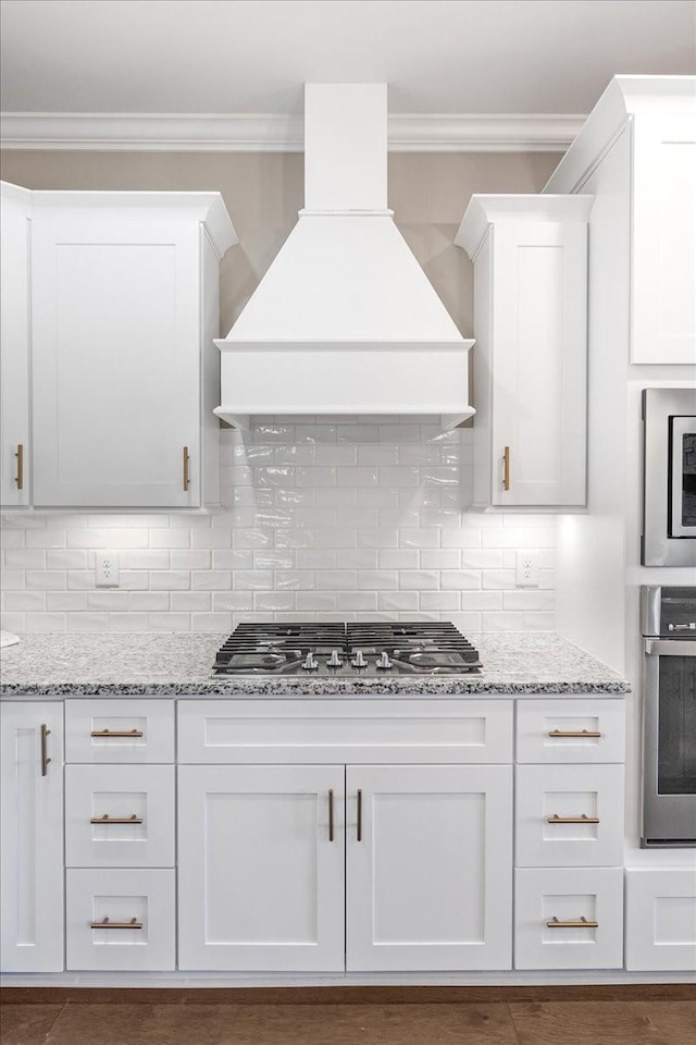 kitchen with custom exhaust hood, white cabinets, crown molding, decorative backsplash, and appliances with stainless steel finishes