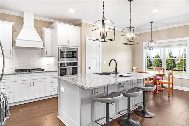 kitchen featuring custom exhaust hood, stainless steel appliances, sink, white cabinetry, and an island with sink