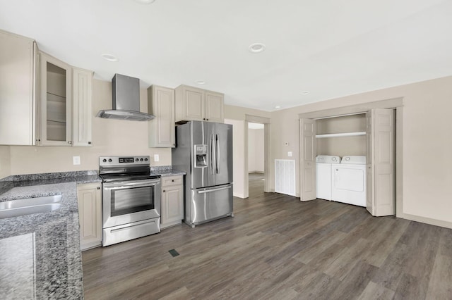 kitchen featuring appliances with stainless steel finishes, dark stone countertops, wall chimney range hood, washing machine and clothes dryer, and dark hardwood / wood-style floors