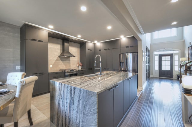 kitchen featuring sink, backsplash, stainless steel appliances, a center island with sink, and wall chimney exhaust hood