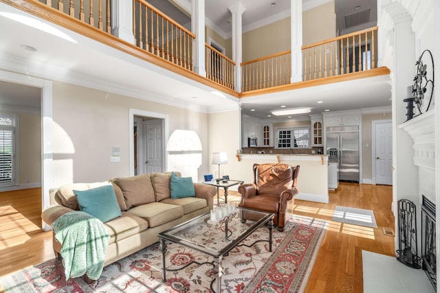 living room featuring a towering ceiling, ornamental molding, a fireplace with flush hearth, light wood-style floors, and ornate columns