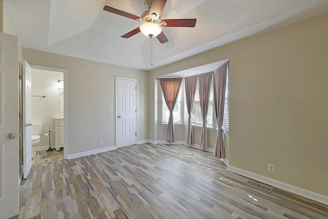 unfurnished bedroom with ensuite bathroom, ceiling fan, a raised ceiling, a textured ceiling, and light hardwood / wood-style flooring