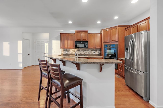 kitchen featuring stone counters, black appliances, a center island with sink, light wood-type flooring, and a kitchen bar