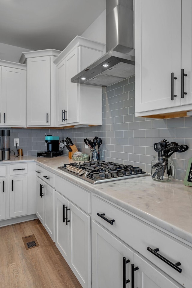 kitchen featuring stainless steel gas stovetop, white cabinets, wall chimney range hood, decorative backsplash, and light wood-type flooring