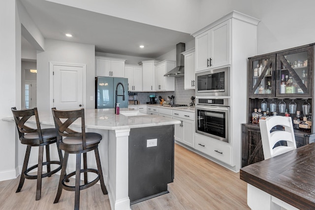 kitchen with wall chimney range hood, tasteful backsplash, a center island with sink, white cabinets, and appliances with stainless steel finishes