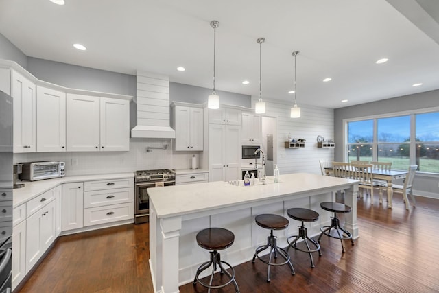 kitchen featuring custom exhaust hood, a kitchen island with sink, stainless steel gas range, decorative light fixtures, and white cabinetry