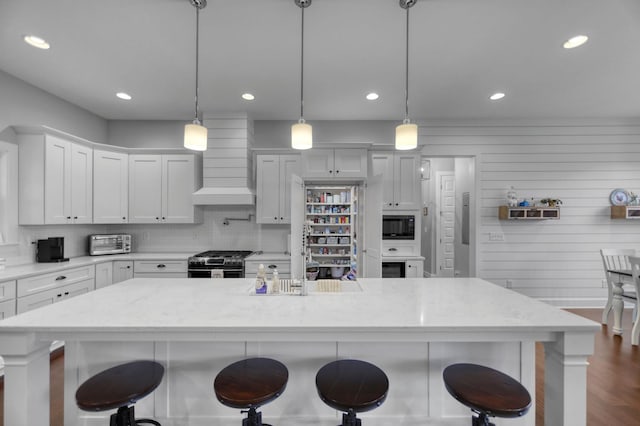 kitchen featuring pendant lighting, stainless steel gas range oven, premium range hood, white cabinetry, and black microwave