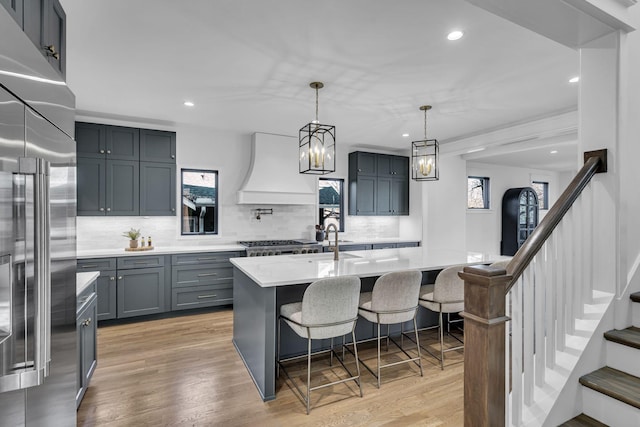 kitchen featuring decorative backsplash, custom range hood, stainless steel appliances, light hardwood / wood-style floors, and an island with sink