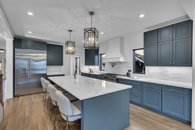 kitchen featuring light stone counters, custom range hood, a center island with sink, high quality appliances, and hanging light fixtures