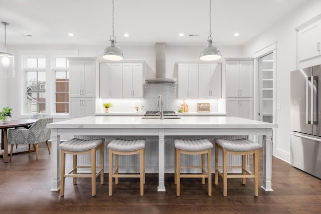 kitchen with stainless steel fridge, wall chimney range hood, white cabinets, hanging light fixtures, and an island with sink