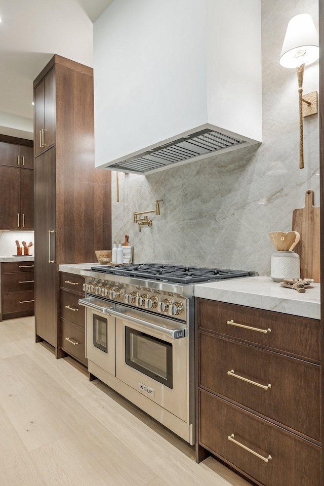 kitchen featuring tasteful backsplash, premium range hood, double oven range, light hardwood / wood-style floors, and dark brown cabinets