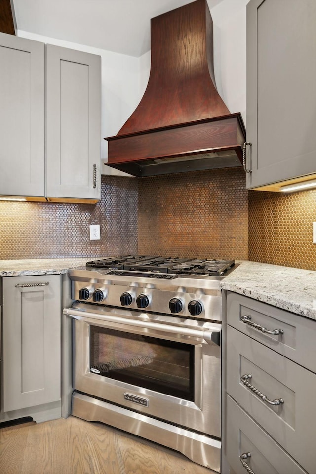 kitchen with decorative backsplash, light stone countertops, gray cabinetry, custom range hood, and stainless steel stove