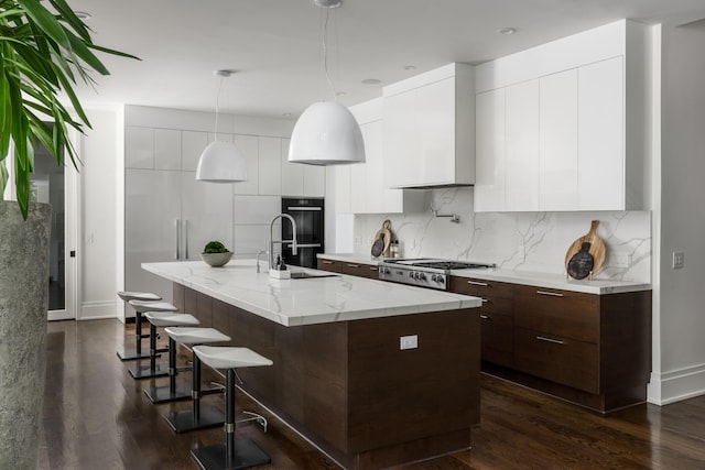 kitchen featuring double oven, a kitchen island with sink, stainless steel gas cooktop, pendant lighting, and white cabinetry