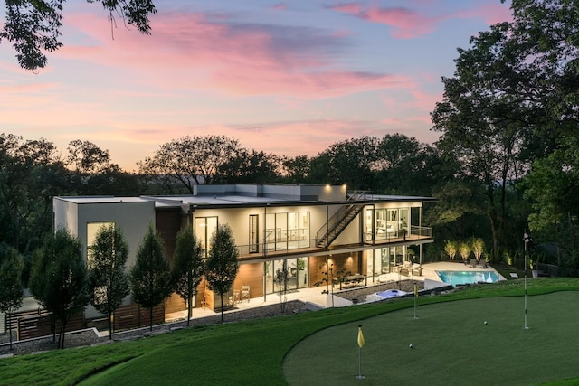 back house at dusk with a lawn, a patio area, a balcony, and a fenced in pool