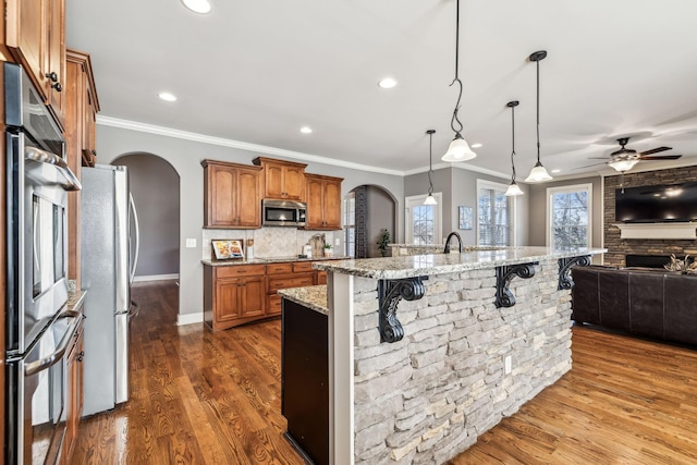 kitchen with ceiling fan, stainless steel appliances, light stone counters, pendant lighting, and a kitchen island with sink