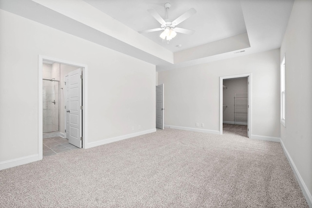 unfurnished bedroom featuring ensuite bathroom, a spacious closet, light carpet, a tray ceiling, and ceiling fan