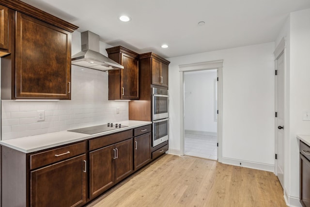 kitchen with wall chimney exhaust hood, stainless steel double oven, light hardwood / wood-style floors, black electric stovetop, and dark brown cabinets