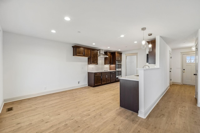 kitchen with dark brown cabinetry, wall chimney range hood, light hardwood / wood-style flooring, double oven, and pendant lighting